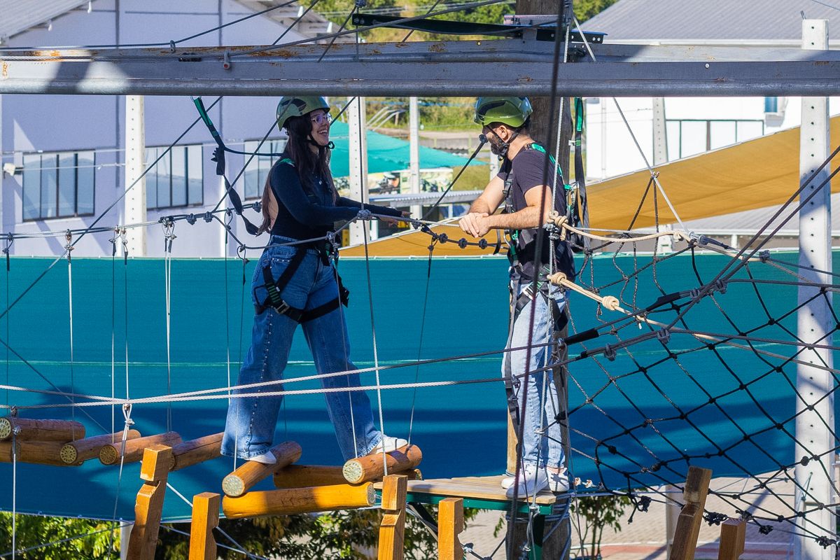 Dois visitantes de capacete e cadeirinha no percurso do Arvorismo Tático